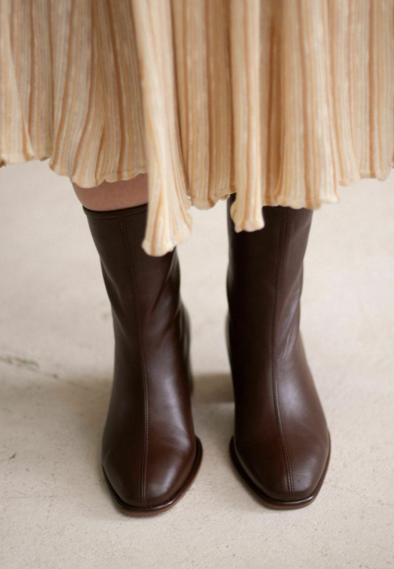 Brown leather ankle boots with a square toe, smooth texture, and vertical stitching, paired with a pleated, light-colored dress.