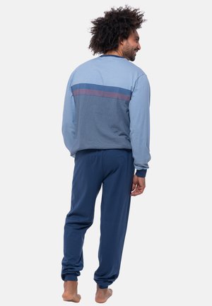 Mann mit lockigem Haar, der ein blau gestreiftes Langarmoberteil und eine einfarbige blaue Hose trägt, steht barfuß mit dem Rücken zur Kamera vor einem einfarbigen Hintergrund.