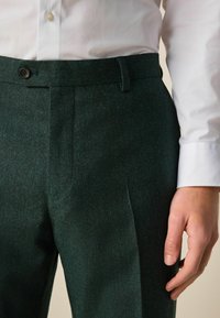 Green wool trousers with a textured finish. Features a flat front, side pockets, and a single button closure at the waist.
