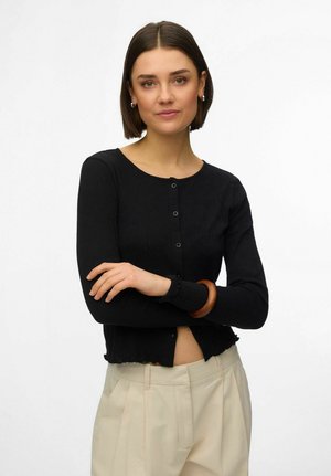 Donna con capelli corti e scuri che indossa un cardigan nero con bottoni, pantaloni a vita alta color avorio e un braccialetto di legno, con le braccia incrociate.