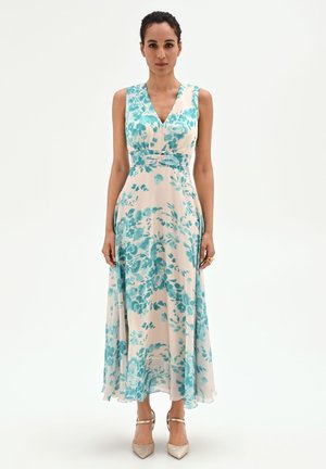 Femme portant une robe blanche sans manches avec motif floral bleu, décolleté en V, taille cintrée et talons beiges à bout pointu, debout sur fond blanc.