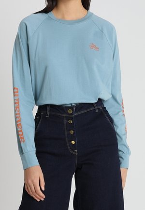 Femme portant une chemise à manches longues bleu clair avec un texte orange sur les manches et la poitrine, associée à un jean taille haute bleu foncé avec des boutons.