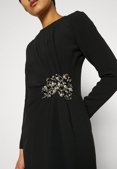 Abito lungo nero a maniche lunghe con tessuto plissettato e un evidente accento di spilla realizzato con vari dettagli metallici e strass in vita.