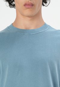 Tricou scurt cu mânecă scurtă, de culoare albastru deschis, realizat din bumbac moale. Prezintă un guler rotund cu cusătură la guler și o croială lejeră.