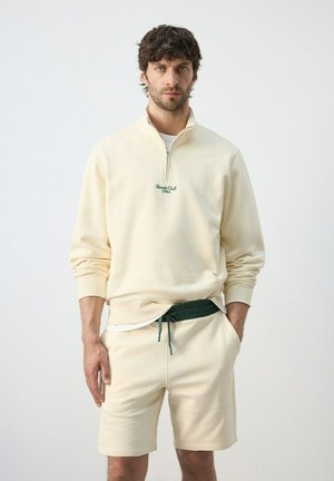 Hombre vistiendo sudadera de color crema con cremallera hasta el pecho y texto "Tennis Club 1984", y pantalones cortos a juego con cordón verde, posando frente a un fondo blanco.
