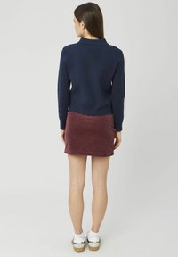 Femme portant un pull marine avec un ourlet festonné, une mini-jupe en velours côtelé bordeaux, des chaussettes blanches et des baskets beige, debout de dos.