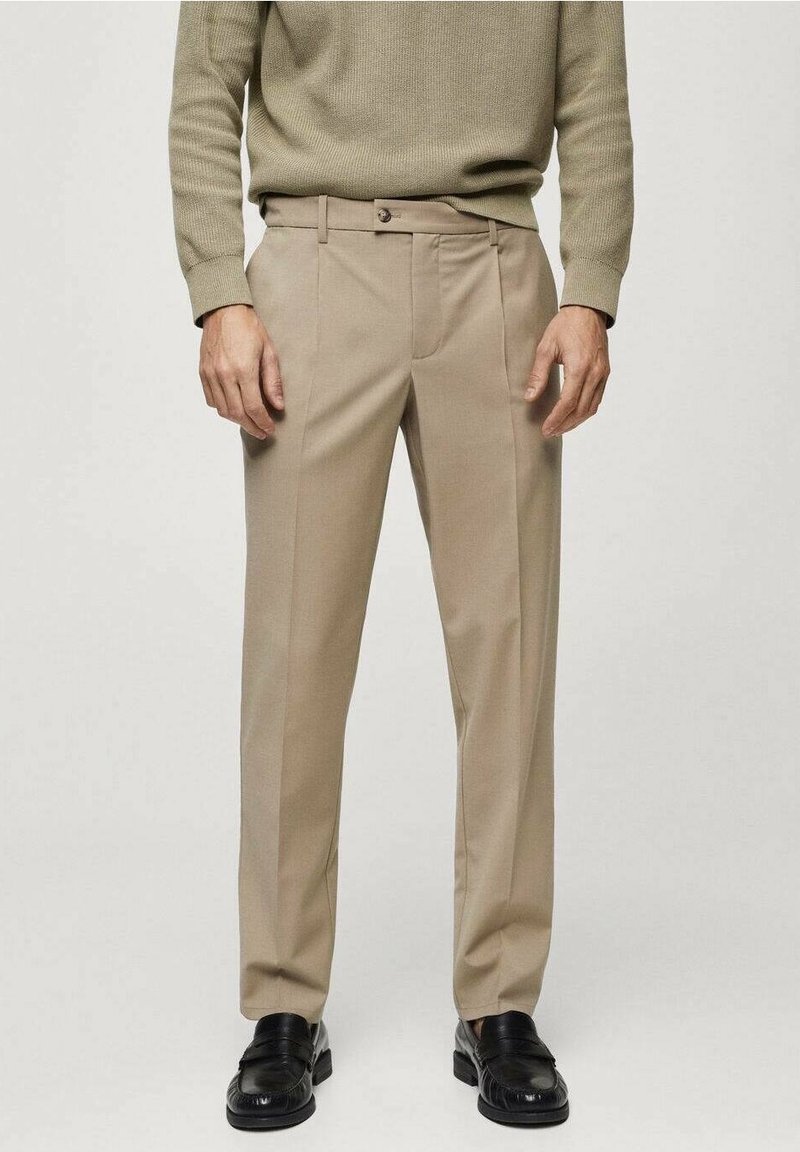 Homme portant un pantalon beige sur mesure, un pull vert olive et des mocassins en cuir noir, debout devant un fond clair uni.