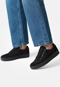 Baskets basses noires en daim et en cuir, avec un dessus texturé, un laçage à l'avant et un accent zip sur le côté.