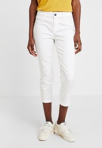 Pantalon blanc court en coton avec une coupe ajustée, doté d'une fermeture à boutons sur le devant et d'un revers roulé, associé à des baskets blanches.