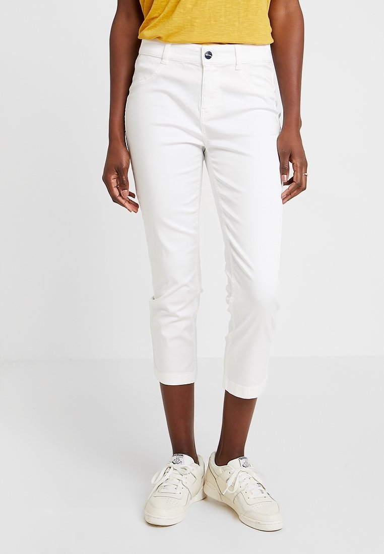 Pantalon blanc court en coton avec une coupe ajustée, doté d'une fermeture à boutons sur le devant et d'un revers roulé, associé à des baskets blanches.