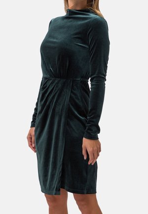 Femme portant une robe en velours vert foncé à manches longues, longueur au genou, avec un col montant et un détail froncé à la taille.