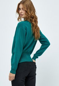 Maglione lavorato a maglia color turchese con una vestibilità comoda, polsini e orlo a coste, presenta un collo alto e una texture liscia. Indossato con pantaloni neri.
