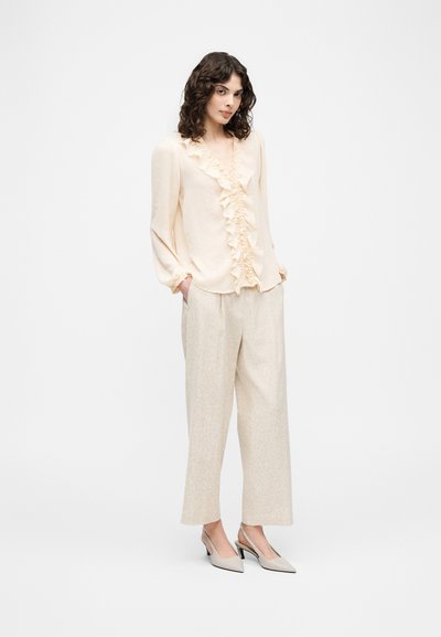 Femme portant un chemisier crème à volants, un pantalon beige à jambes larges et des talons slingback beiges, debout les mains dans les poches sur fond blanc.