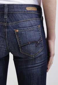 Jeans de mezclilla azul oscuro con corte ajustado, que presentan un parche de cuero con marca, costuras en contraste y un bolsillo con detalle de costura curva.