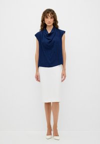 Top senza maniche blu navy con scollatura drappeggiata e motivo testurizzato, abbinato a una gonna bianca al ginocchio e tacchi bianchi.