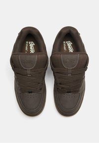 Hnedé tenisky s hladkými a textúrovanými povrchmi, vybavené zaoblenou špičkou, hrubými šnúrkami a logom na jazyku a boku.