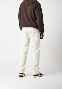 Pantalon cargo blanc à coupe décontractée, avec poches latérales et poches arrière, associé à un sweat à capuche marron et des baskets foncées.