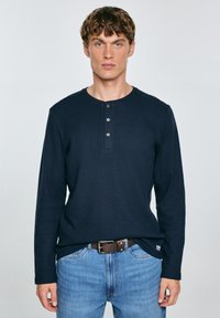 Maglietta henley a maniche lunghe blu navy con una texture a coste, dotata di una patta con bottoni e una vestibilità rilassata, abbinata a jeans azzurri.