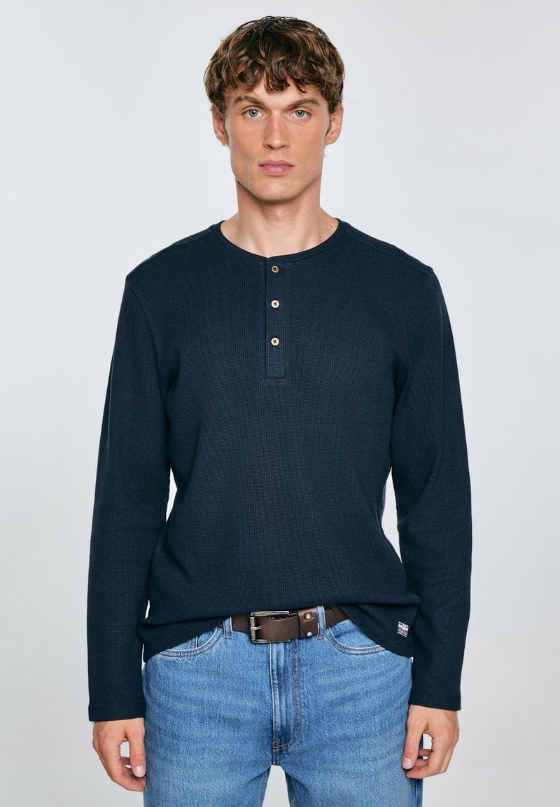 Maglietta henley a maniche lunghe blu navy con una texture a coste, dotata di una patta con bottoni e una vestibilità rilassata, abbinata a jeans azzurri.