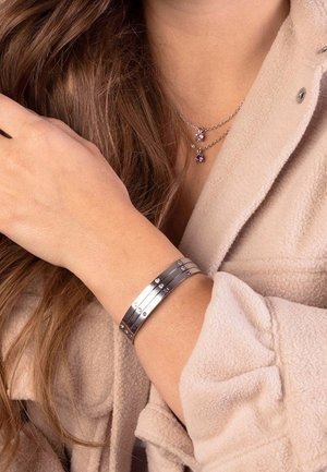 Bracelet manchette en argent présentant des bandes horizontales à finition polie et des petites pierres claires dispersées. Porté au poignet avec un tissu texturé.