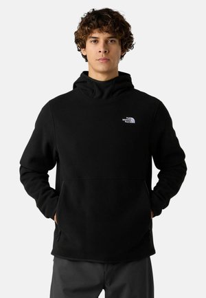 Giovane uomo con capelli ricci che indossa un felpa con cappuccio nera di The North Face, con le mani nelle tasche anteriori, in posa frontale su uno sfondo semplice.