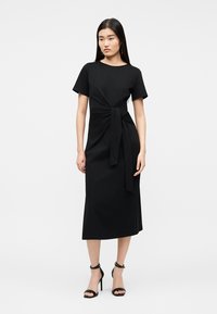 Ženska, ki stoji, oblečena v črno midi obleko s kratkimi rokavi in detajlom sprednje zavezave, kombinirano s črnimi sandali z visokimi petami.