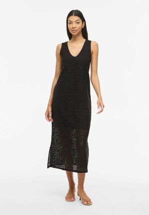 Femme portant une robe midi noire en crochet sans manches avec un col en V et des sandales beiges à talons ouverts, debout devant un fond blanc uni.