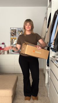 Camiseta marrón y pantalones ajustados negros, sosteniendo una caja de Zalando. La habitación cuenta con estanterías con ropa y revistas en el fondo.