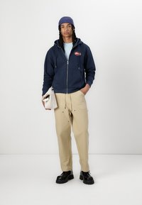 Marinblå zip-up hoodie, ljusblå skjorta, beige byxor, svarta klumpiga skor. Modellen håller en beige väska och bär en blå mössa.