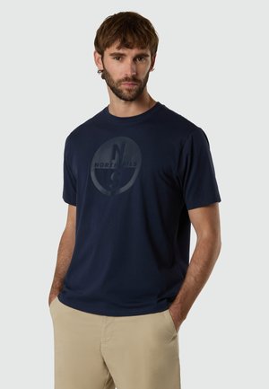 Uomo che indossa una maglietta North Sails blu navy e pantaloni beige, in piedi con le mani nelle tasche davanti a uno sfondo grigio chiaro uniforme.