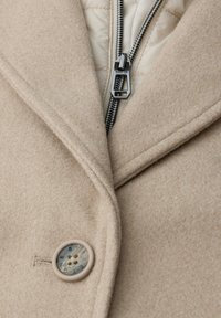 Beige uldblandingsfrakke med en stor revers, en lynlås lukning og en dekorativ knap med en struktureret overflade. Inderforet har en quiltet foring.