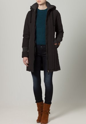 Schwarze gesteppte Jacke mit Kapuze, mit grauen gerippten Bündchen, einem Reißverschluss und seitlichen Taschen. Kombiniert mit einem türkisfarbenen gestrickten Pullover und dunklen Jeans.