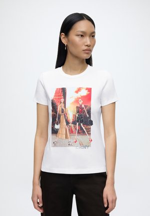 Femme aux longs cheveux noirs portant des boucles d'oreilles en or en forme d'anneau, t-shirt blanc avec un graphique de deux illustrations de mode marchant dans un paysage urbain.