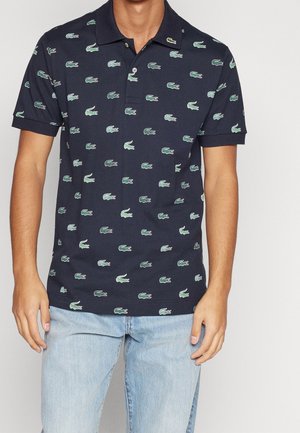 Hombre con camiseta polo azul marino con logotipos repetidos de cocodrilo verde y pantalones vaqueros azul claro, de pie sobre fondo liso.