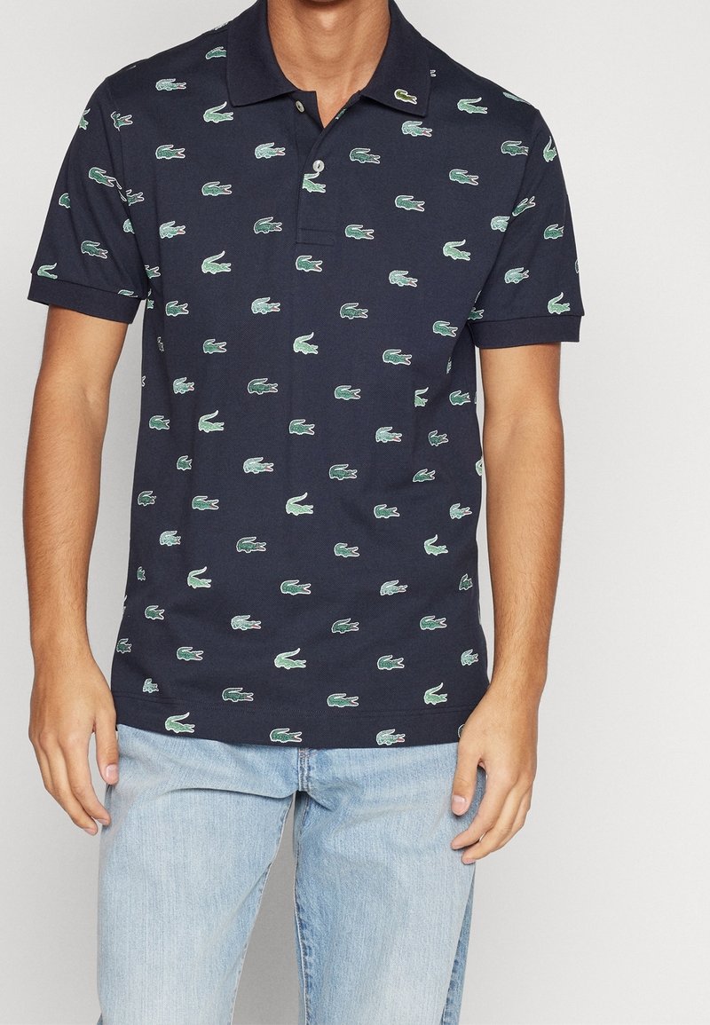Homme portant un polo bleu marine avec des logos de crocodile verts répétés et un jean bleu clair, debout devant un fond uni.