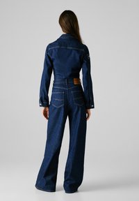 Combinaison en denim bleu marine avec manches longues, taille cintrée, jambes évasées et couture contrastante. Présente une étiquette en patch au dos.
