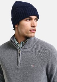 Grauer gerippter Pullover mit einem Viertel-Reißverschluss-Kragen, über einem blauen und weißen Karohemd getragen, kombiniert mit einer navyblauen gerippten Strickmütze.