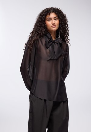 Czarna, przezroczysta bluzka z dużym kołnierzem w kształcie kokardy, długimi rękawami i luźnym krojem, połączona z luźnymi czarnymi spodniami o gładkiej fakturze.