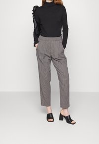 Woman wearing black turtleneck, checked high-waist trousers, black heeled mules, and carrying a black ruched shoulder bag.