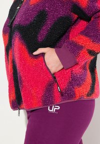 Veste en polaire floue multicolore avec des motifs vibrants rouges, violets et noirs, dotée d'une poche zippée. Pantalons de sport violets avec un logo blanc.