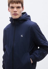 Μπλε ναυτικό μπουφάν με κουκούλα από ελαφρύ ύφασμα, με φερμουάρ, ελαστικές μανσέτες και μικρό λογότυπο στο πάνω αριστερό μέρος του στήθους.