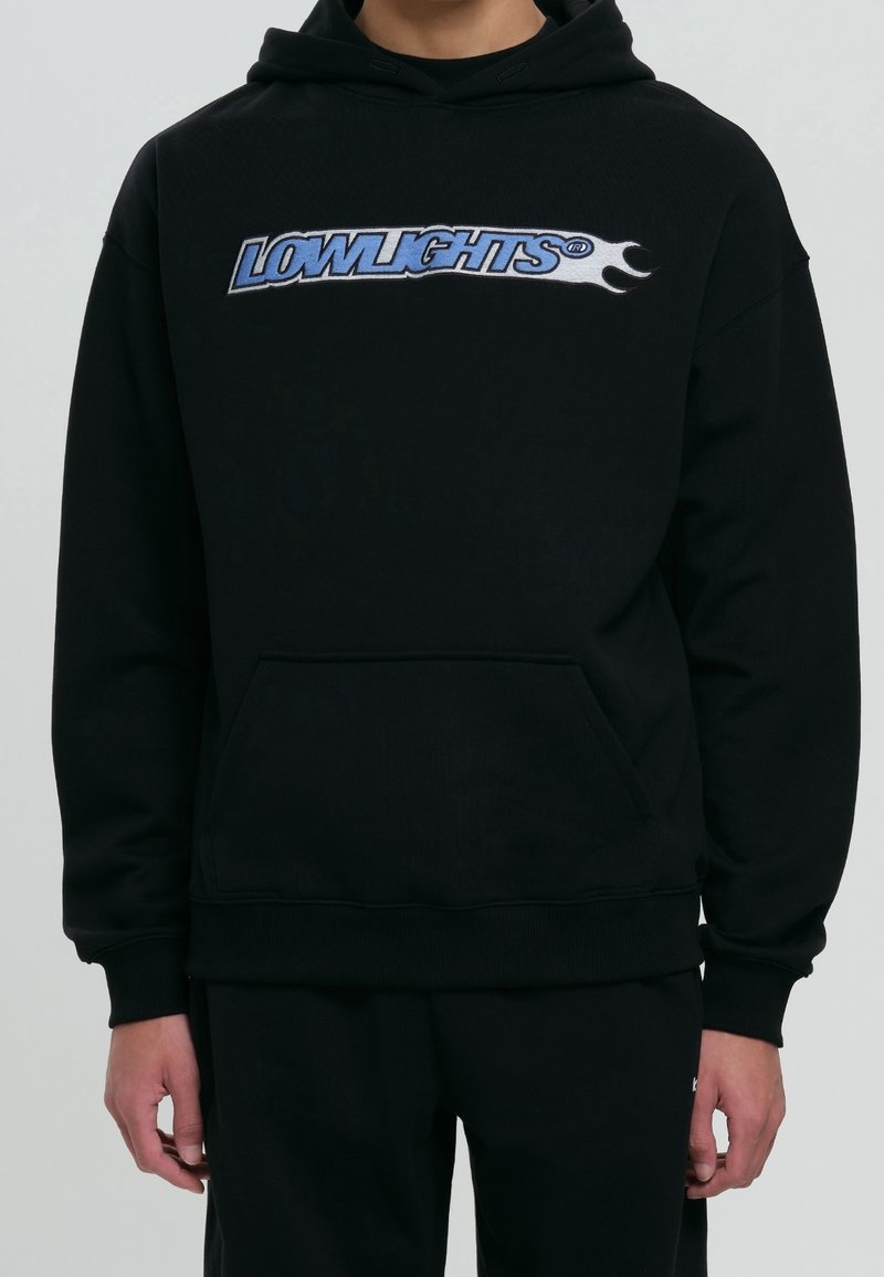 Personne portant un sweat à capuche noir avec le texte "LOWLIGHTS" en bleu et blanc ainsi qu'un motif de flammes sur la poitrine, les mains détendues le long du corps.