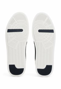 Semelles blanches de baskets avec un motif de bande de roulement texturé et des patchs noirs au talon, avec la marque "Tommy Hilfiger" au centre de chaque semelle.
