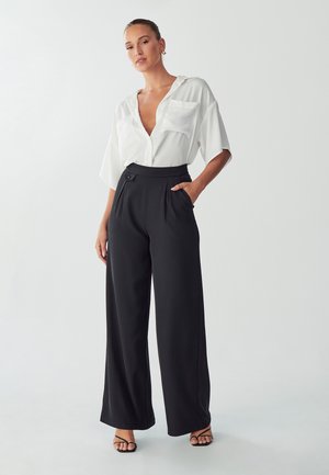 White short-sleeve blouse with a V-neck and two chest pockets, paired with high-waisted black wide-leg trousers featuring a button and side pockets.