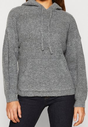 Femme portant un sweat à capuche en maille grise avec une poche avant et un pantalon foncé, cadrée au niveau du cou et des cuisses.