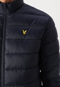 Marineblå puffjakke med quilted tekstur, glidelås og en gull brodert ørn logo på brystet. Lett og isolert design.