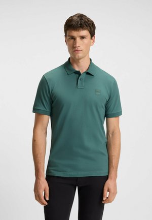 Chemise polo verte en tissu lisse, dotée d'un col, d'une patte de boutonnage à deux boutons et d'un petit logo sur la poitrine. Manches courtes.