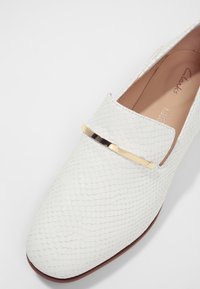 Mocassin blanc texturé avec un détail en barre métallique dorée sur le dessus et une semelle intérieure beige marquée "Clarks."