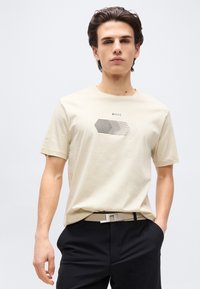 Νέος άνδρας με σκούρα μαλλιά, που φοράει μπεζ μπλουζάκι με σχέδιο, μαύρο παντελόνι και μπεζ ζώνη με γεωμετρική αγκράφα, στέκεται μπροστά σε λευκό φόντο.