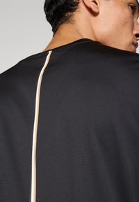 Melns sporta krekls ar teksturētu audumu un centrālu vertikālu šuvi, ko izceļ plānas baltas un zelta līnijas.